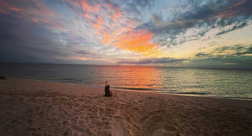 The sky appears in gentle colors above the beach. A person is sitting on the beach with their hand raised into the air. 